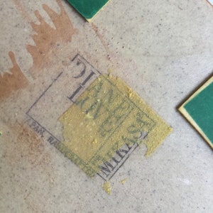 Op de afbeelding: Close-up van een tan gekleurde keramische tegel met een gouden sticker met de tekst "Mikasa, Japan" en een groene sticker in de rechterbovenhoek.
