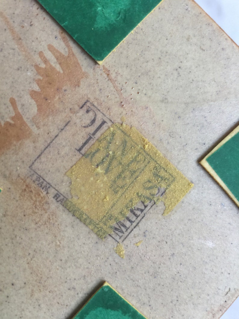 Op de afbeelding: Close-up van een tan gekleurde keramische tegel met een gouden sticker met de tekst "Mikasa, Japan" en een groene sticker in de rechterbovenhoek.