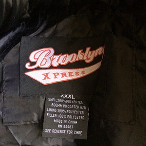 Puede incluir: Una chaqueta negra con un parche de logotipo Brooklyn XPRESS rojo y blanco. La etiqueta indica que la chaqueta es XXXL, hecha de 100% poli&eacute;ster y fabricada en China. La etiqueta tambi&eacute;n incluye instrucciones de cuidado.