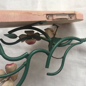 May include: A pink wooden shelf with a green metal floral design. The shelf is attached to the wall with screws and a metal bracket.