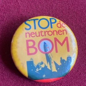 May include: A round yellow pinback button featuring blue and pink text that reads STOP de neutronen BOM. The design includes a graphic illustration of a dark blue rocket silhouette rising above a stylized cityscape against the yellow background.