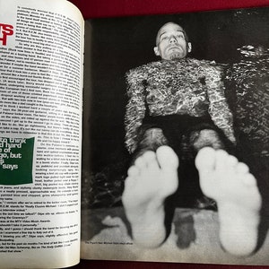 May include: A black and white photo of a man submerged in water. The man is wearing a dark shirt and shorts. His feet are visible above the waterline. The caption below the photo reads "The Pixies' best Michael Price stays afloat."
