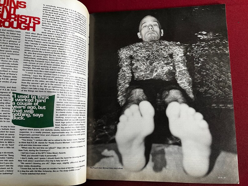 May include: A black and white photo of a man submerged in water. The man is wearing a dark shirt and shorts. His feet are visible above the waterline. The caption below the photo reads "The Pixies' best Michael Price stays afloat."