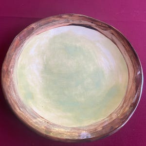 May include: Hand-painted ceramic plate with a light green center and a brown rim. The plate has a slightly irregular shape, adding to its handmade charm. The plate is a decorative item.