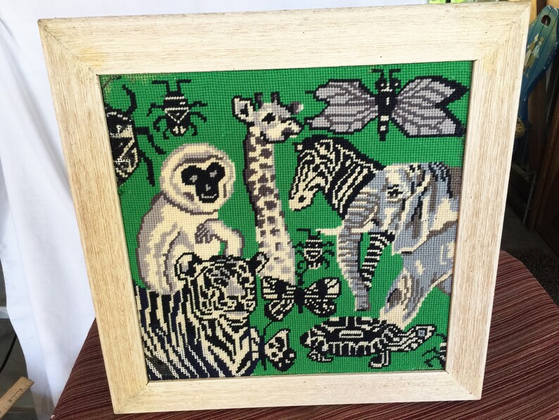 May include: A framed needlepoint picture with a green background featuring a variety of animals including a zebra, giraffe, elephant, monkey, tiger, and butterflies.