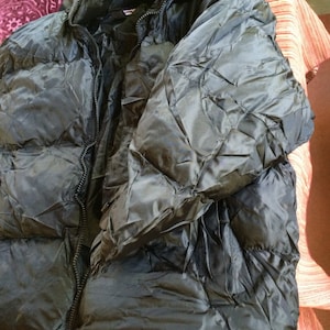 Puede incluir: Una chaqueta acolchada negra con cierre de cremallera y capucha. La chaqueta tiene un dise&ntilde;o acolchado y est&aacute; hecha de un material brillante y resistente al agua.
