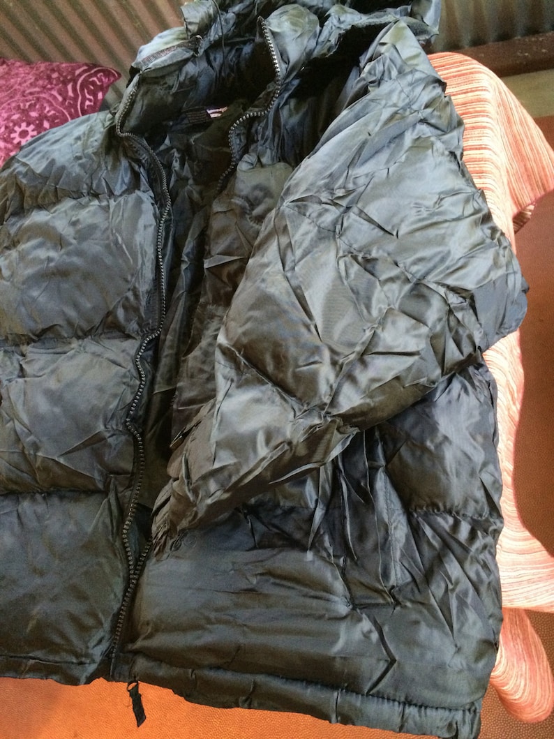 Puede incluir: Una chaqueta acolchada negra con cierre de cremallera y capucha. La chaqueta tiene un dise&ntilde;o acolchado y est&aacute; hecha de un material brillante y resistente al agua.