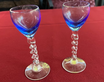 Pode incluir: Dois copos de shot de vidro transparente com borda azul e haste torcida. Os copos estão sobre uma superfície vermelha.