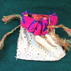 May include: A small white fabric pouch with a pink and blue trim and gold and green tassels. The pouch is decorated with white lace.