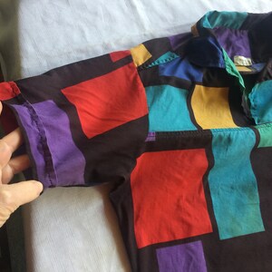 K&ouml;nnte beinhalten: Ein kurz&auml;rmeliges Hemd mit Kn&ouml;pfen und einem geometrischen Muster in Rot-, Blau-, Gelb-, Lila- und Gr&uuml;nt&ouml;nen auf schwarzem Hintergrund.