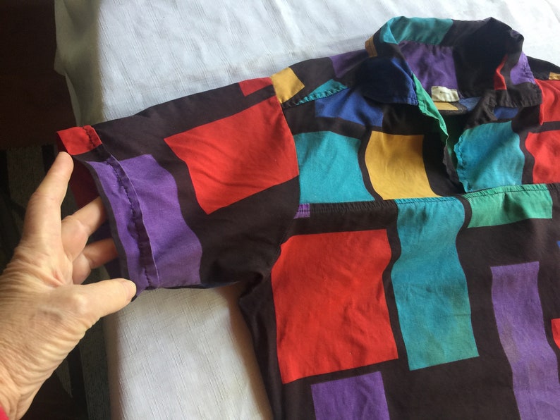 K&ouml;nnte beinhalten: Ein kurz&auml;rmeliges Hemd mit Kn&ouml;pfen und einem geometrischen Muster in Rot-, Blau-, Gelb-, Lila- und Gr&uuml;nt&ouml;nen auf schwarzem Hintergrund.