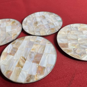 May include: Four round coasters made of white and brown mother of pearl tiles. The tiles are arranged in a checkered pattern. The coasters have a silver metal rim.