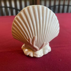 May include: A ceramic seashell ornament in off-white with pink detailing. The shell features a ribbed design and rests on a textured base. The ornament is displayed on a red surface.