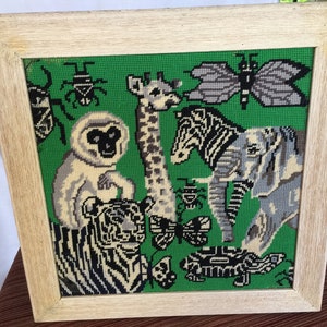 May include: A framed needlepoint picture with a green background featuring a variety of animals including a tiger, zebra, giraffe, monkey, elephant, and butterfly. The animals are all stitched in black, white, and gray thread.