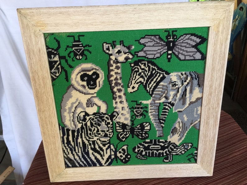 May include: A framed needlepoint picture with a green background featuring a variety of animals including a tiger, zebra, giraffe, monkey, elephant, and butterfly. The animals are all stitched in black, white, and gray thread.