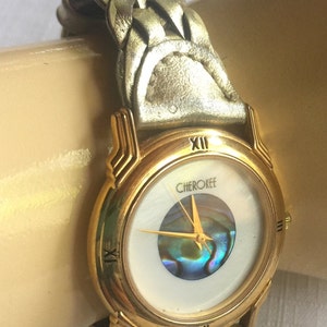 May include: Gold-tone wristwatch with a white face and a braided leather band. The watch face features a mother-of-pearl inlay and the word "Cherokee" printed on it.