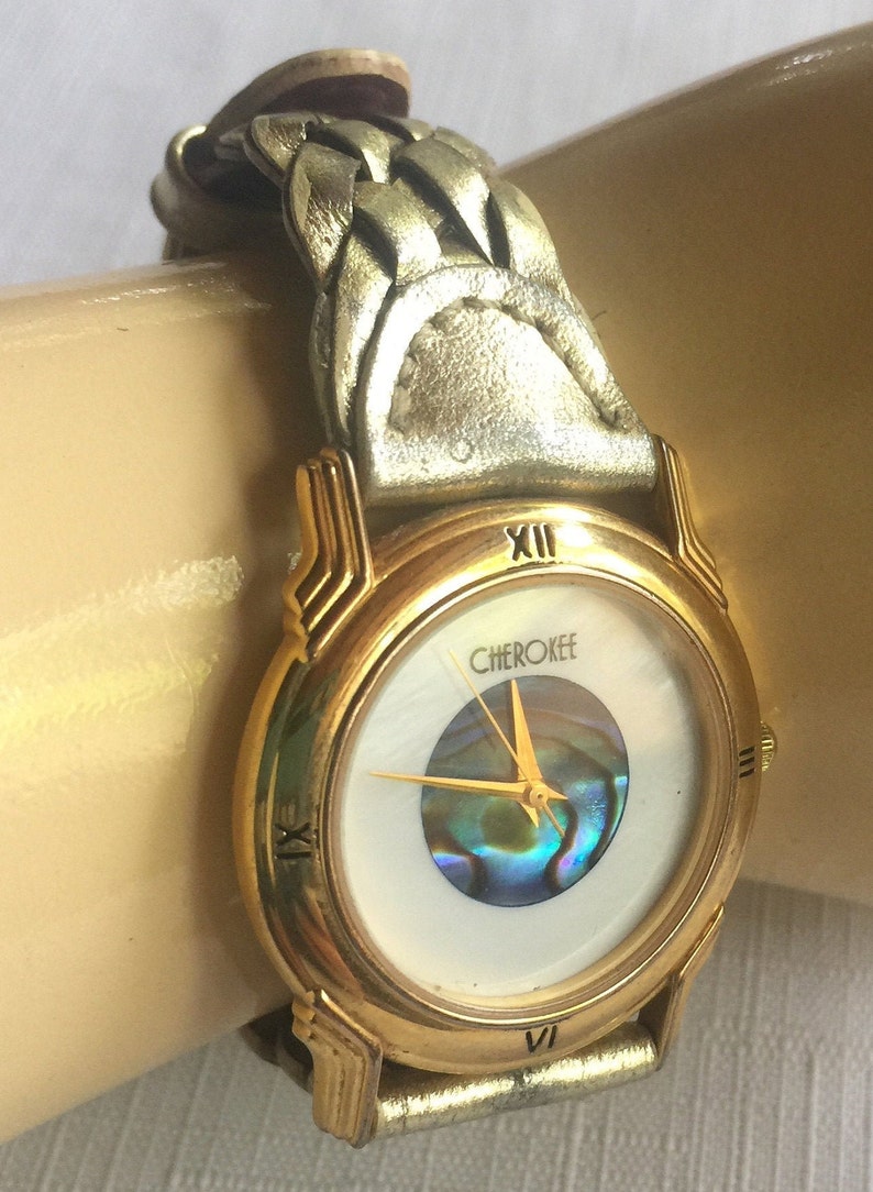 May include: Gold-tone wristwatch with a white face and a braided leather band. The watch face features a mother-of-pearl inlay and the word "Cherokee" printed on it.