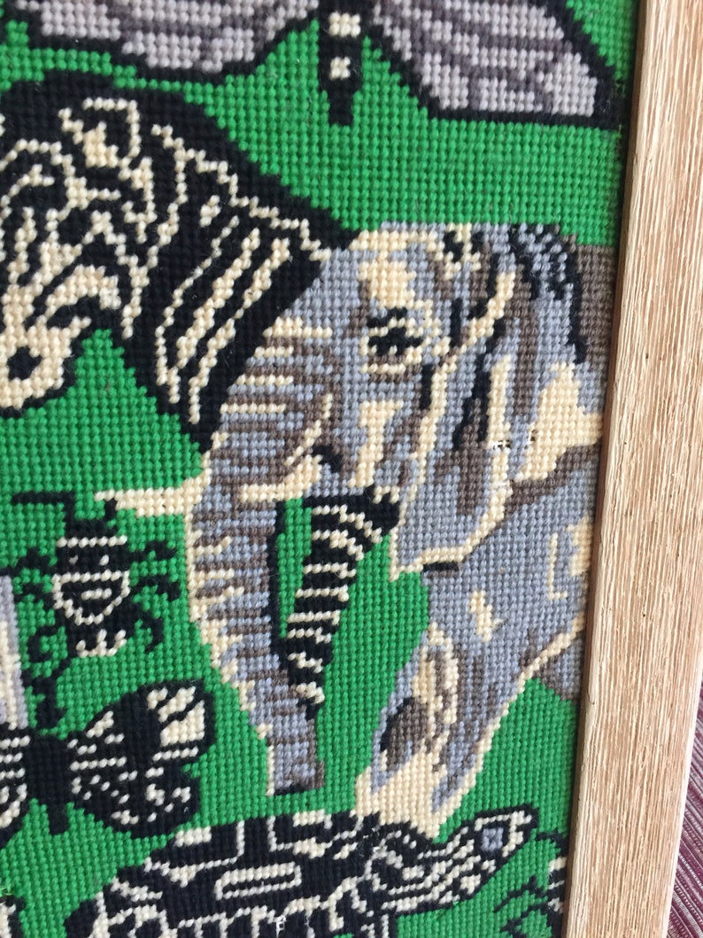 May include: A framed needlepoint tapestry with a green background and a variety of animals including a zebra, an elephant, and a turtle. The animals are stitched in various shades of gray, black, and white.