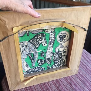 May include: A framed tapestry with a green background and a black and white design of animals, including a zebra, a monkey, and a giraffe. The tapestry is in a wooden frame with a brown finish.