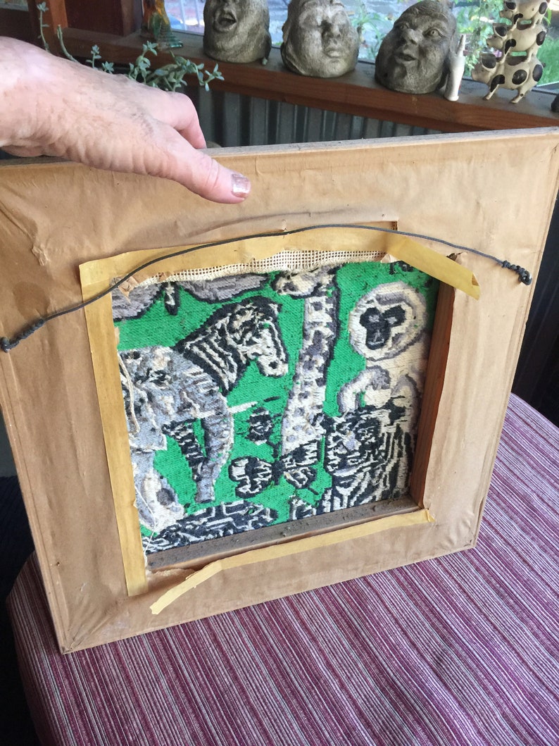 May include: A framed tapestry with a green background and a black and white design of animals, including a zebra, a monkey, and a giraffe. The tapestry is in a wooden frame with a brown finish.