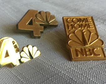 Puede incluir: Tres pines de color dorado con el logotipo del pavo real de NBC. Un pin tiene el número "4" y las palabras "Come Home to" sobre el logotipo. Otro pin tiene el número "4" y un logotipo de pavo real más pequeño. El tercer pin tiene las palabras "Come Home to" y "NBC" debajo del logotipo.