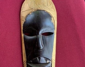 May include: A wooden mask with a black face and gold accents. The mask has a carved design on the forehead and around the eyes. The mouth is open and has a carved design.