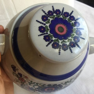 Op de afbeelding: Een witte theepot van keramiek met een blauw en bloemmotief. De deksel heeft een grote blauwe bloem met roze en groene accenten.