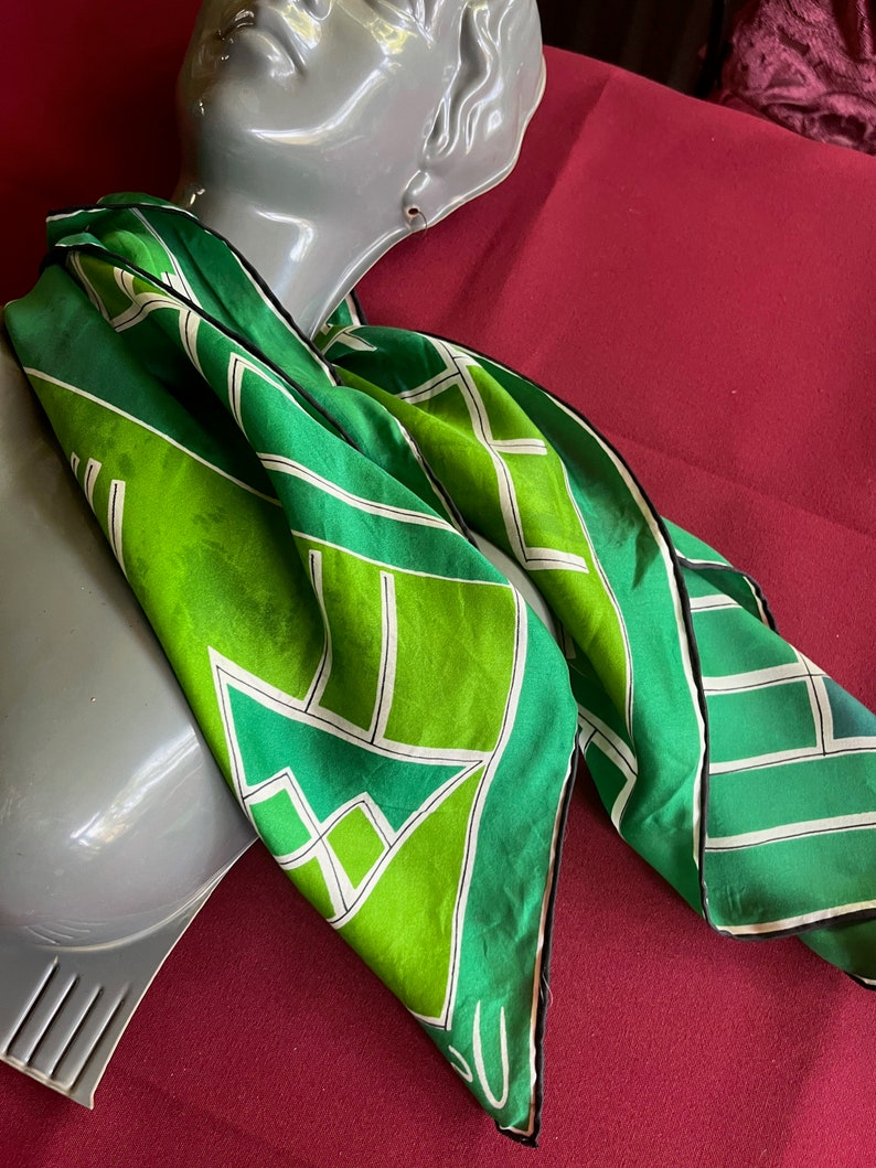 May include: A green and white silk scarf with a geometric pattern. The scarf is folded and draped over a mannequin's neck.