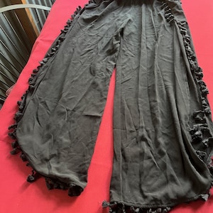Puede incluir: Pantalones negros de pierna ancha con ribete de volantes y borlas. Los pantalones están hechos de una tela ligera y tienen una cintura elástica. Las borlas están a lo largo de las costuras exteriores y en la parte inferior de las piernas.