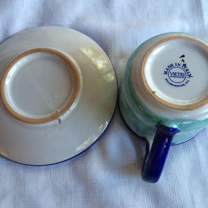 May include: A blue and green ceramic teacup and saucer set with a blue handle. The saucer has a blue rim. The teacup has a blue rim and a white circle with the text "Made in Italy" and "Vietri Incorporated, Hillsborough, NC".