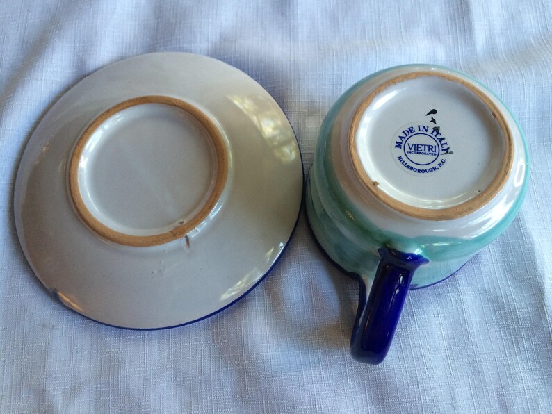 May include: A blue and green ceramic teacup and saucer set with a blue handle. The saucer has a blue rim. The teacup has a blue rim and a white circle with the text "Made in Italy" and "Vietri Incorporated, Hillsborough, NC".
