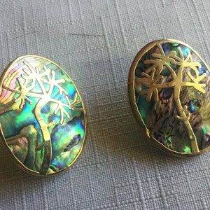 May include: Two oval-shaped earrings with a gold-colored metal frame. The earrings feature an iridescent abalone shell inlay with a gold tree design. The abalone shell displays shades of green, blue, and purple.
