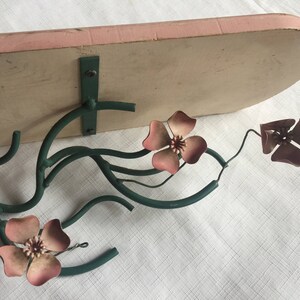 May include: A metal wall shelf with a pink painted top and a green metal floral design. The shelf has three pink flowers with five petals each.