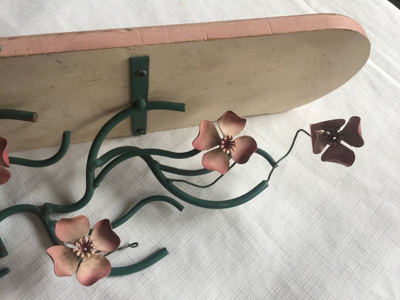 May include: A metal wall shelf with a pink painted top and a green metal floral design. The shelf has three pink flowers with five petals each.