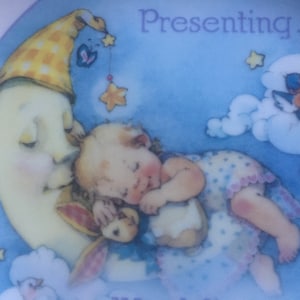 Pode incluir: Um beb&ecirc; dormindo em uma lua crescente com um chap&eacute;u xadrez amarelo e branco. A lua est&aacute; cercada por nuvens e estrelas. O texto "Presenting..." est&aacute; no topo da imagem.