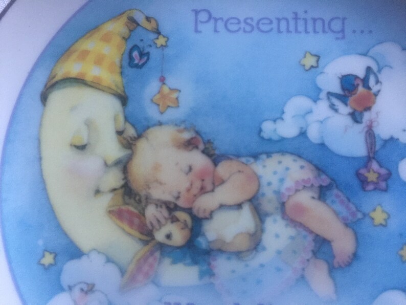 Pode incluir: Um beb&ecirc; dormindo em uma lua crescente com um chap&eacute;u xadrez amarelo e branco. A lua est&aacute; cercada por nuvens e estrelas. O texto "Presenting..." est&aacute; no topo da imagem.