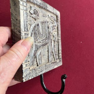 May include: A square, wooden wall hook with a carved elephant design. The hook is painted in shades of gray and white, with a black metal hook. The elephant carving is detailed and the hook is attached to the bottom of the square.
