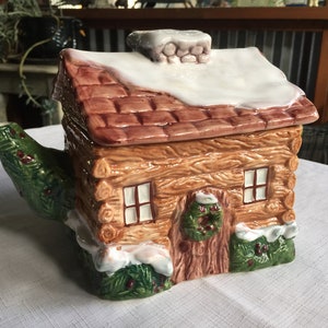 Può includere: Una teiera in ceramica a forma di baita con tetto innevato, rivestimento in legno marrone e una ghirlanda verde sulla porta. La teiera ha un beccuccio verde e marrone a forma di pino.
