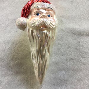 以下が含まれることがあります： サンタクロースの頭の形をしたクリスマスオーナメント。白いグリッターのトリムが付いた赤い帽子、青い目、グリッターのアクセントが付いた長い白いひげがあります。上部には吊り下げ用の金色の金属ループが付いています。