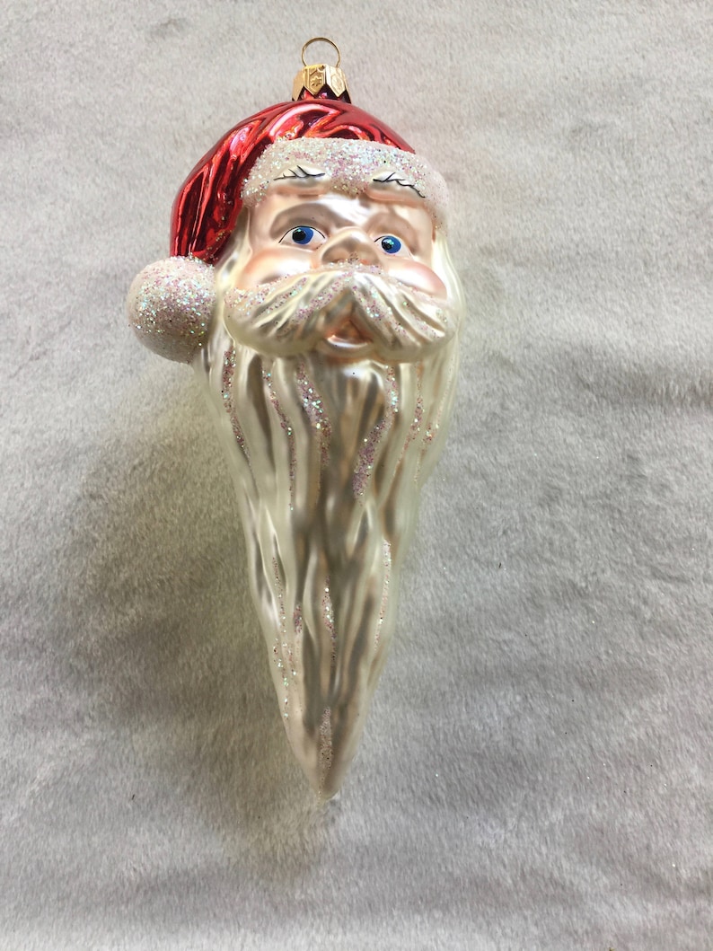 以下が含まれることがあります： サンタクロースの頭の形をしたクリスマスオーナメント。白いグリッターのトリムが付いた赤い帽子、青い目、グリッターのアクセントが付いた長い白いひげがあります。上部には吊り下げ用の金色の金属ループが付いています。