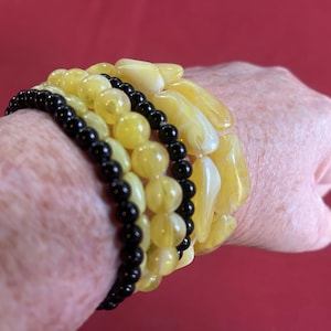 May include: A collection of bracelets in shades of yellow and black. The bracelets include round beads and irregular, polished stones. The background is a solid red.