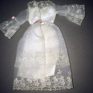 Puede incluir: Un vestido blanco de gasa con detalles de encaje y una cinta de raso rosa alrededor de la cintura.