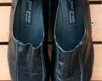 May include: A pair of black leather slip-on shoes with white stitching. The shoes have a suede heel and a label that reads "Paul Green München".