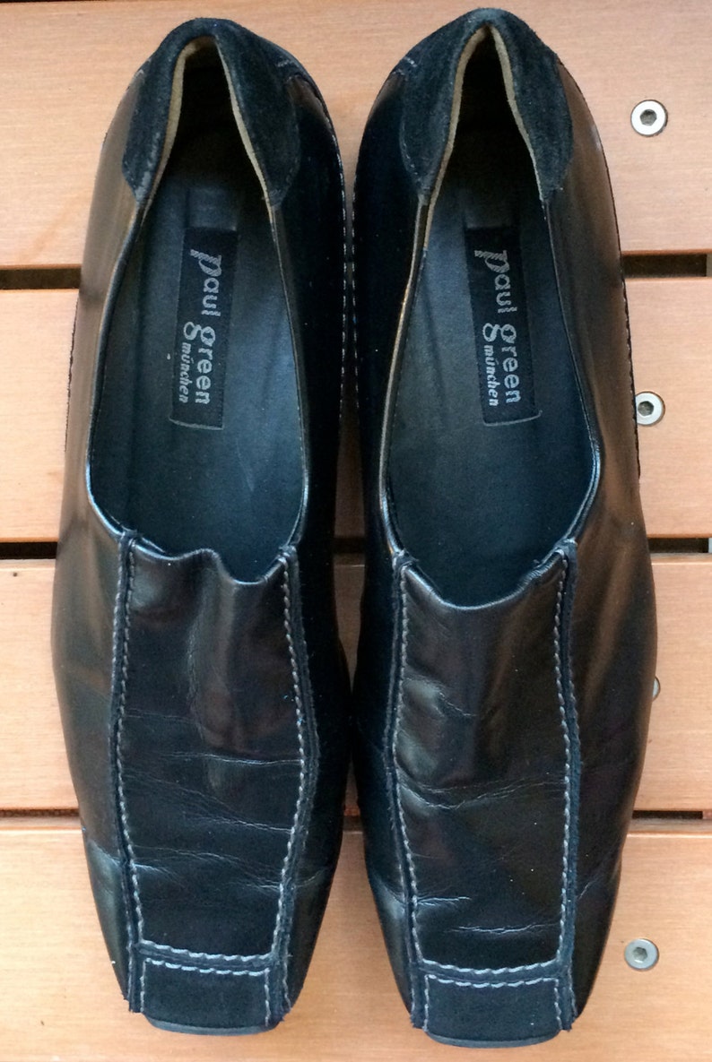 Puede incluir: Un par de zapatos negros de cuero sin cordones con costuras blancas. Los zapatos tienen un tac&oacute;n de ante y una etiqueta que dice "Paul Green M&uuml;nchen".