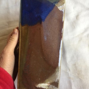 Op de afbeelding: Een keramische vaas met een blauwe, bruine en gouden glazuur. De vaas heeft een rechthoekige vorm met een licht afgeschuinde bovenkant.