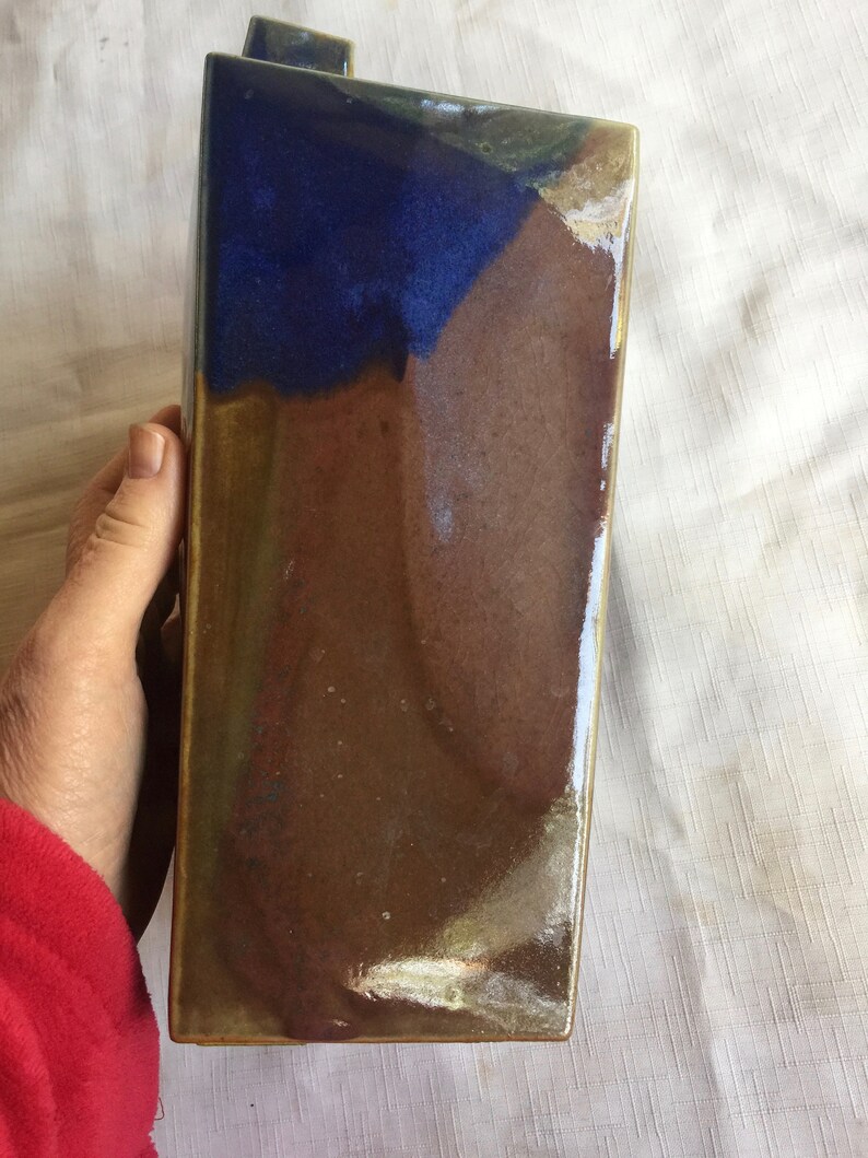 Op de afbeelding: Een keramische vaas met een blauwe, bruine en gouden glazuur. De vaas heeft een rechthoekige vorm met een licht afgeschuinde bovenkant.