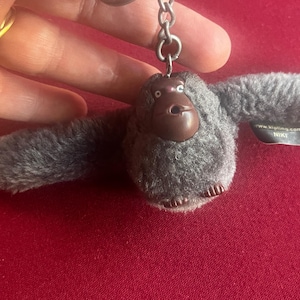 Peut inclure: Un porte-clés gorille en peluche grise avec un visage marron et les bras tendus. Le porte-clés a un anneau en métal argenté et une petite étiquette qui dit "www.kipling.com NIKI".