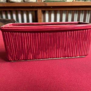 Pode incluir: Uma floreira retangular de cerâmica vermelha com um design texturizado de ranhuras verticais. A floreira está sobre uma superfície vermelha. O fundo inclui uma prateleira de madeira e metal ondulado.