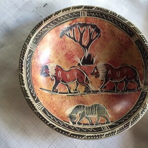 May include: A round, brown, stone bowl with a black and white geometric design around the rim. The bowl features a painted scene of two lions walking and a zebra grazing in a savanna setting with a tree in the background.