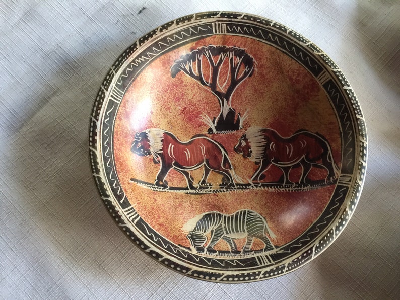 May include: A round, brown, stone bowl with a black and white geometric design around the rim. The bowl features a painted scene of two lions walking and a zebra grazing in a savanna setting with a tree in the background.